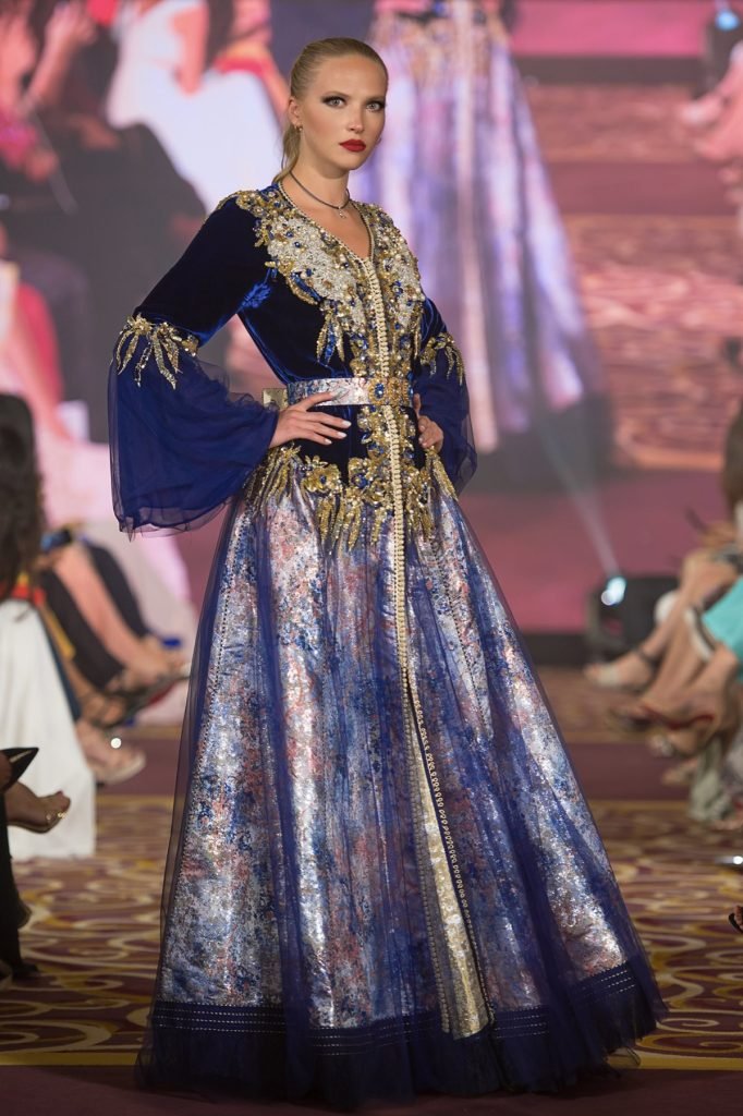 Une mannequin défile avec élégance sur le podium, vêtue d’une somptueuse tekchita mauve ornée de broderies raffinées.